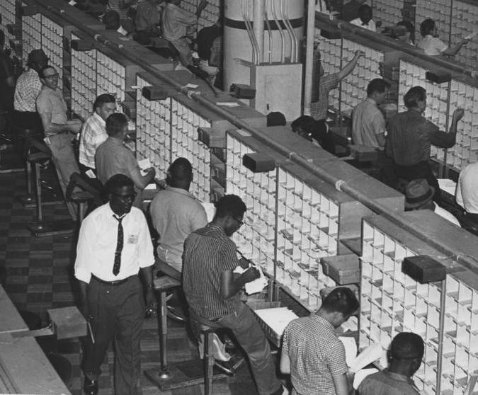 Detail of photograph of postal clerks -- both black and white -- sorting mail at the Washington, D.C., Post Office circa 1958, with a black supervisor walking in the foreground.